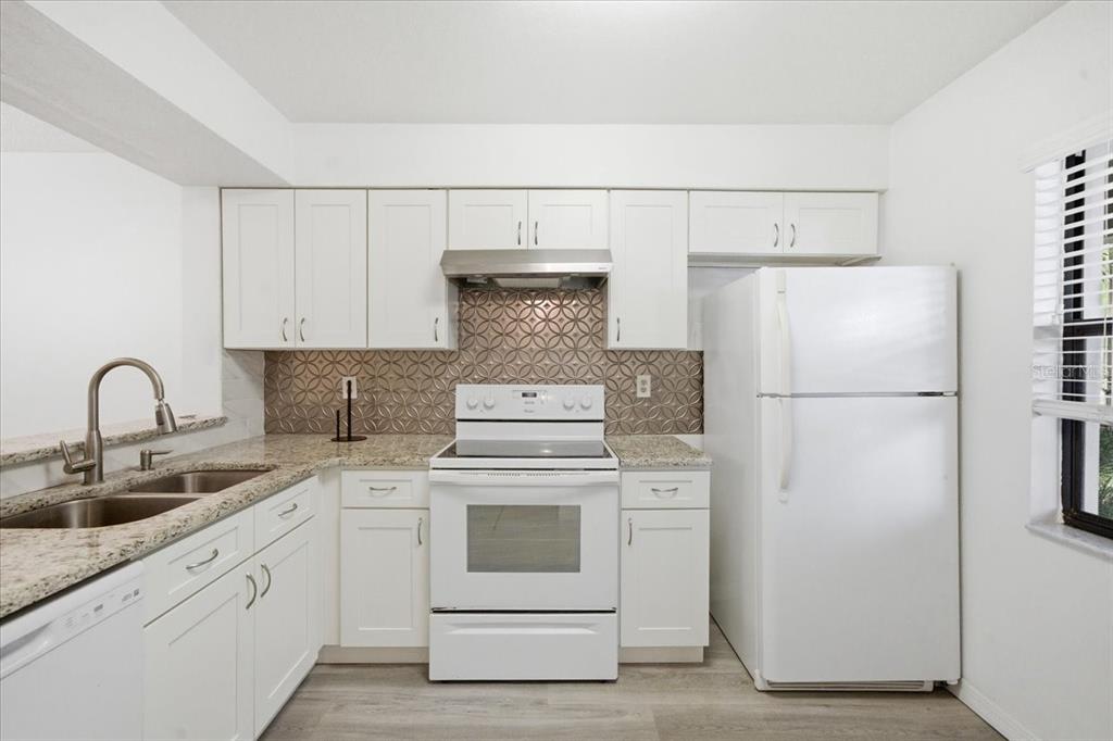 A kitchen features white cabinets, granite countertops, a patterned backsplash, and white appliances — 3399 MERMOOR DRIVE UNIT 101, PALM HARBOR, FL
