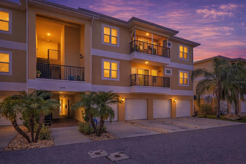 A three-story apartment building exterior features tan stucco walls, private balconies, and ground-level garages — 6395 BANYAN BOULEVARD UNIT 303, NEW PORT RICHEY, FL
