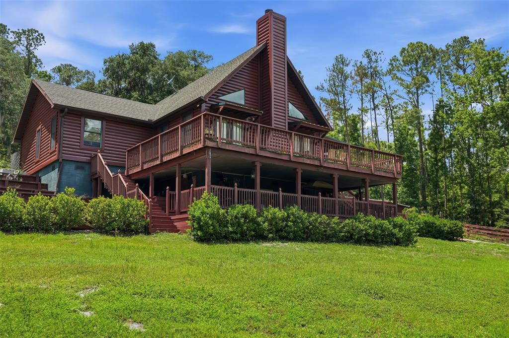 A two-story log cabin with a large wrap-around wooden deck sits on a grassy hill — 24050 WINDING CREEK DRIVE, BROOKSVILLE, FL