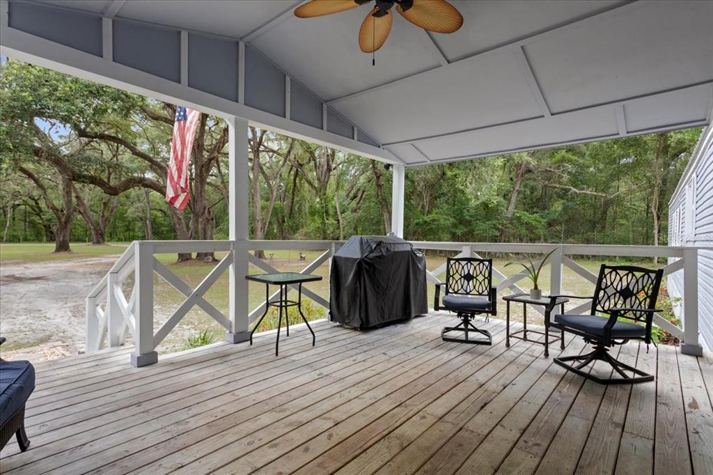 A covered wooden deck features a ceiling fan, outdoor furniture, and a grill overlooking trees — 16221 SW 67TH PLACE, ARCHER, FL