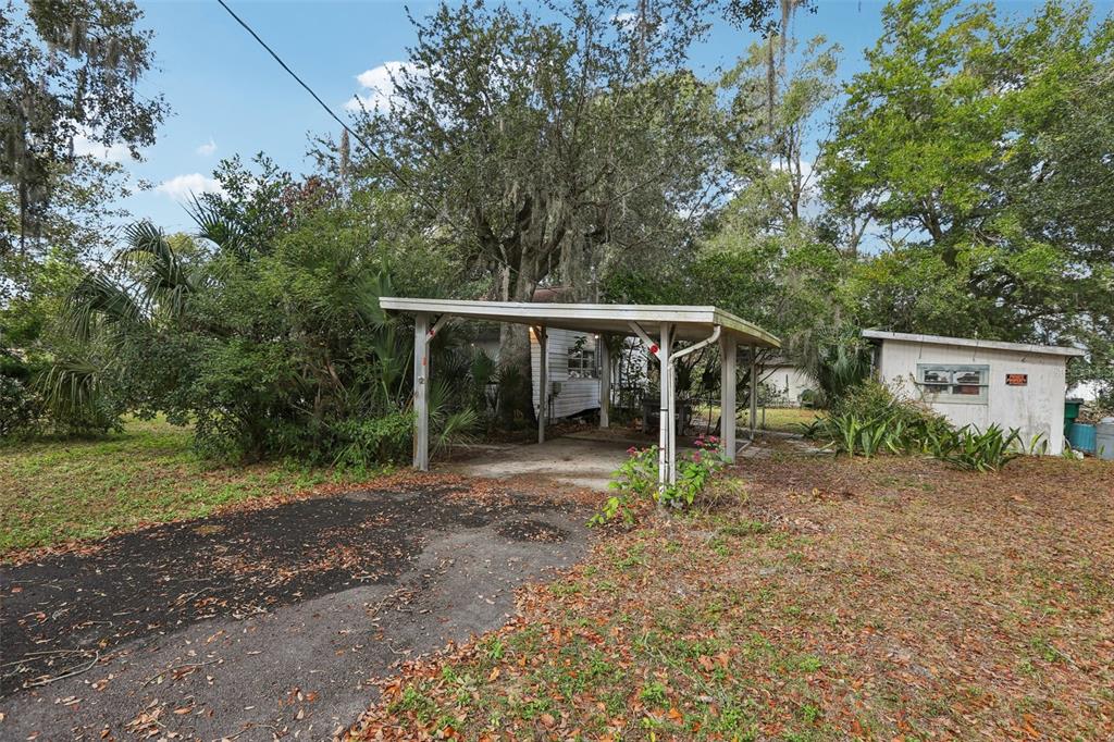 A gravel driveway leads to a metal carport beside a small, white residential building — 38025 13TH AVENUE, ZEPHYRHILLS, FL