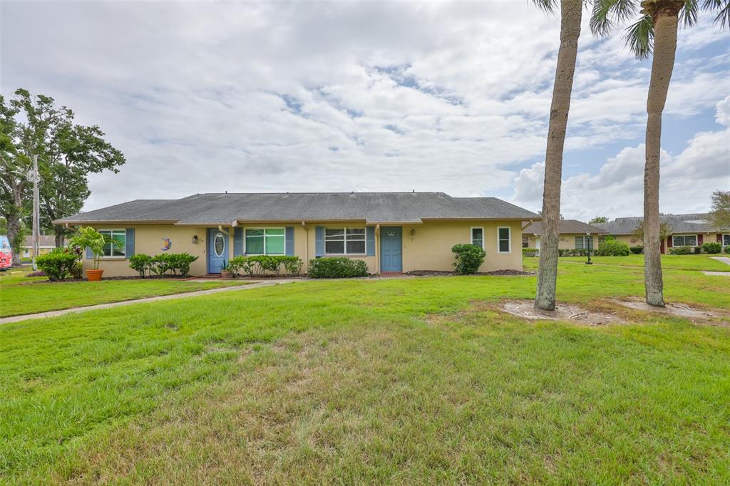 A single-story tan duplex with blue doors and a gray roof sits on a lawn — 1908 CANTERBURY LANE UNIT 20, SUN CITY CENTER, FL