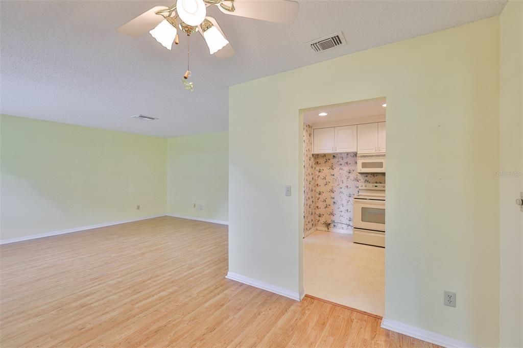 A light-walled living room with wood-look flooring opens into a kitchen with floral wallpaper — 1908 CANTERBURY LANE UNIT 20, SUN CITY CENTER, FL