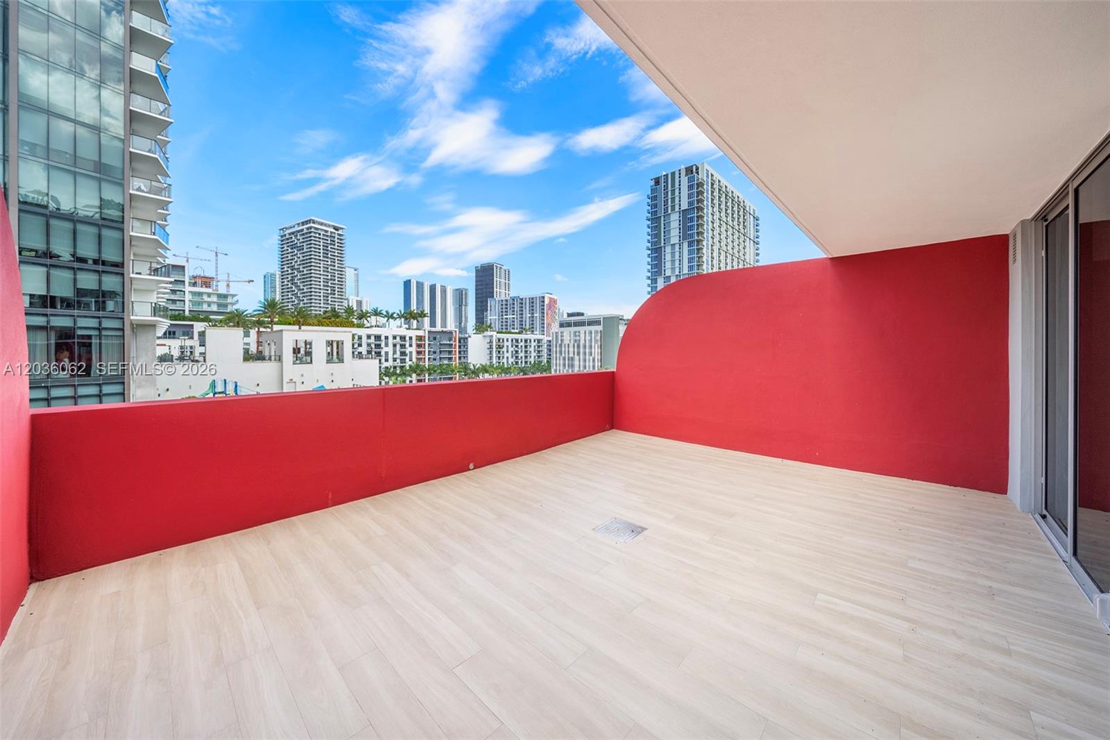 A spacious balcony features light wood-look flooring, a vibrant red wall, and city skyline views — MIAMI, FL