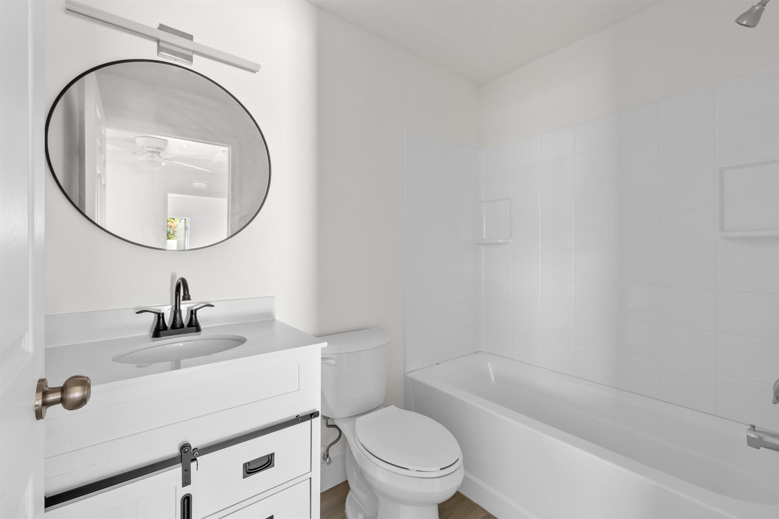 A bathroom features a white vanity, round mirror, toilet, and a white tiled tub surround — 1135 17th Place, Vero Beach FL 32960, VERO BEACH, FL