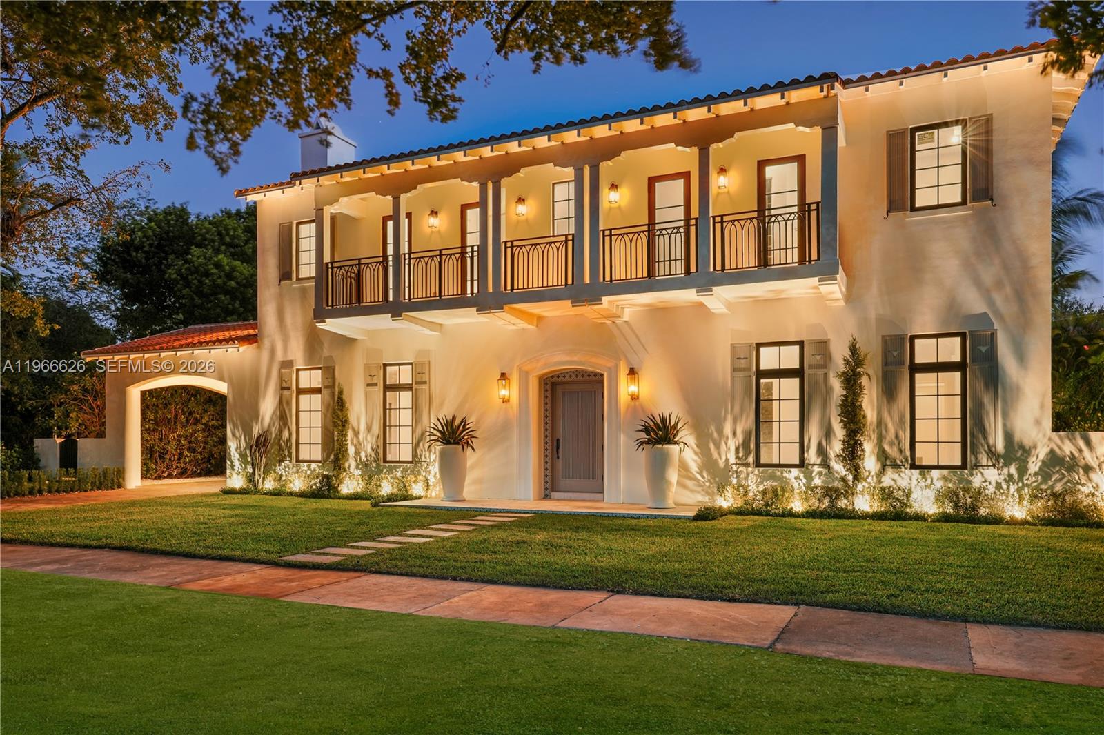 A two-story Mediterranean-style house with a balcony, arched entryway, and exterior landscape lighting at dusk — 1514 Consolata Ave, Coral Gables FL 33146, CORAL GABLES, FL