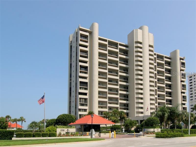 A tall, beige multi-story residential building with a red-roofed gatehouse and an American flag — 1290 GULF BOULEVARD UNIT 307, CLEARWATER, FL