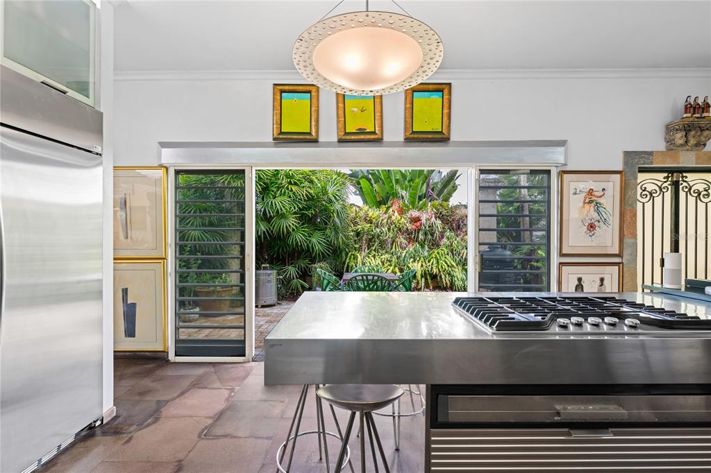 A modern kitchen features a stainless steel island, stone flooring, and sliding doors opening to greenery — 13 PEDROSA ST. GARDEN HILLS, GUAYNABO, PR