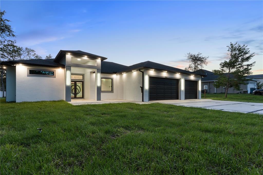 A modern single-story house with a white stucco exterior, black garage doors, and recessed lighting — 9201 NIGHTINGALE ROAD, WEEKI WACHEE, FL