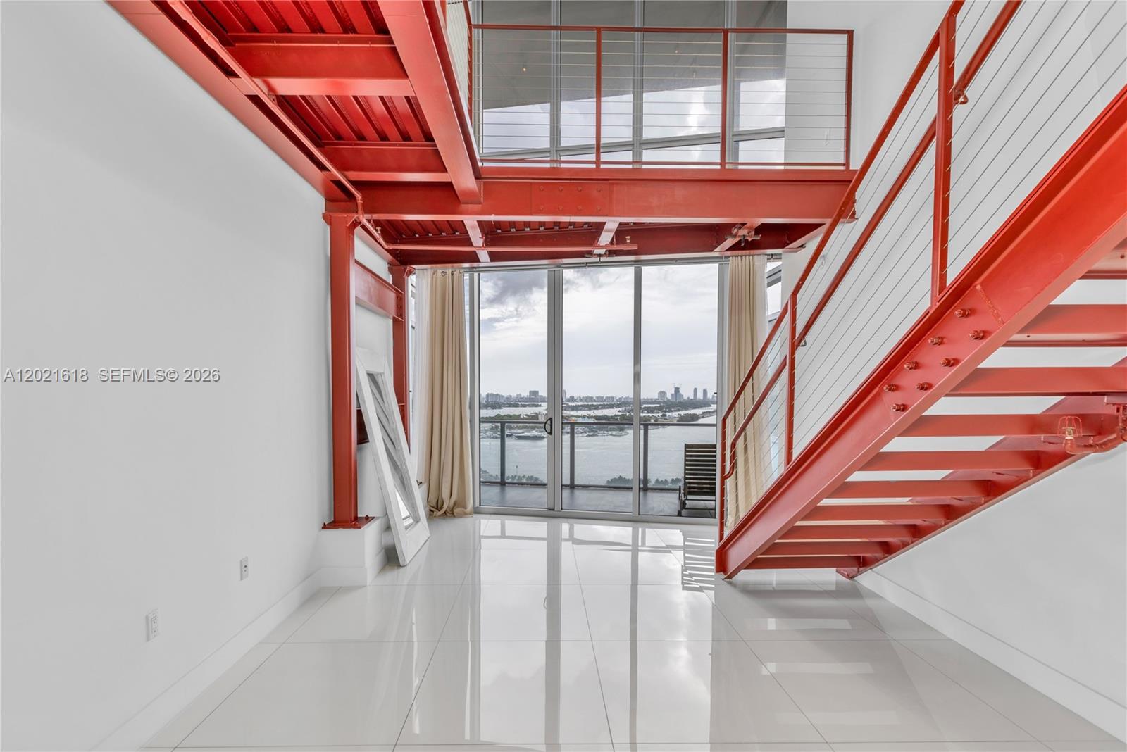 A modern room features red industrial steel beams, a staircase, and floor-to-ceiling windows overlooking water — 1100 Biscayne Blvd # 2706, Miami FL 33132, MIAMI, FL