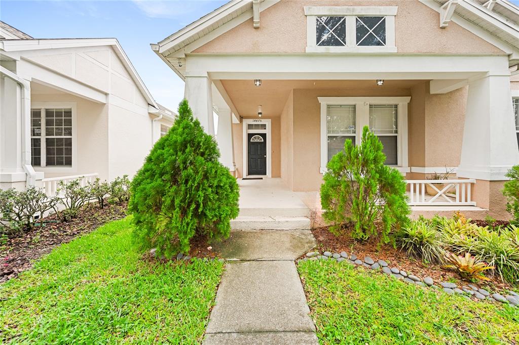 A front view of a tan house with a covered porch and two small evergreen trees — 14475 JAMAICA DOGWOOD DRIVE, ORLANDO, FL
