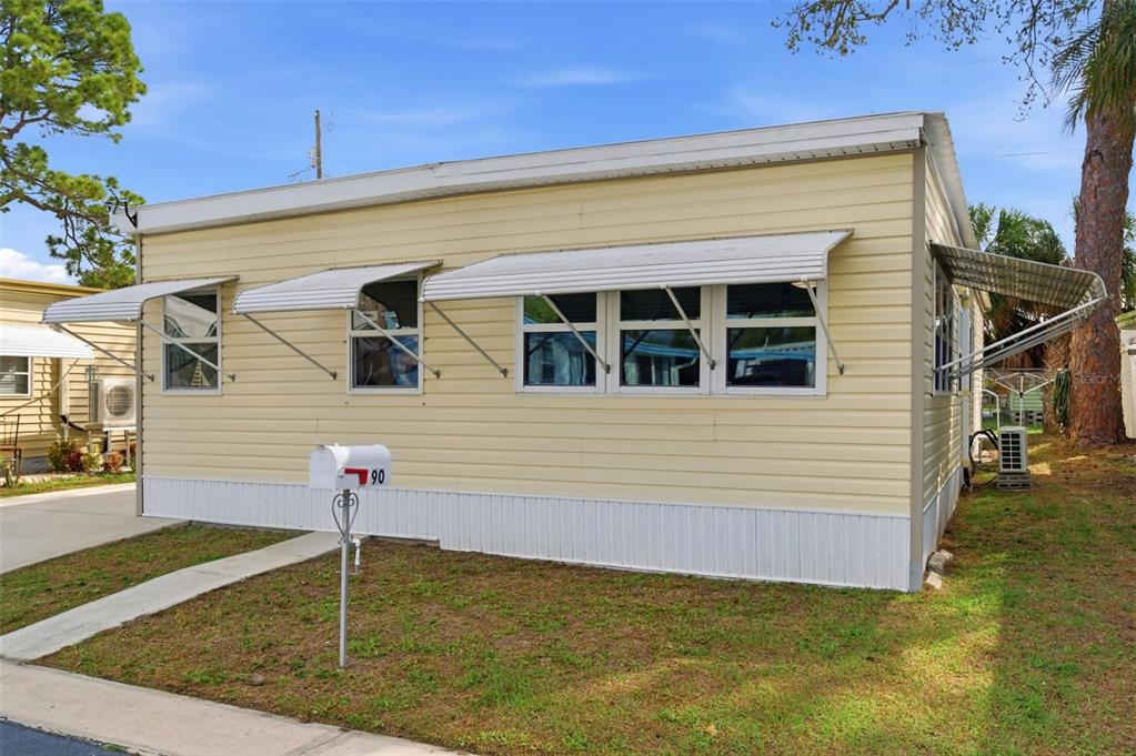 A light yellow mobile home with metal window awnings and a mailbox in the front yard — 7803 46TH AVENUE N UNIT 90, ST PETERSBURG, FL