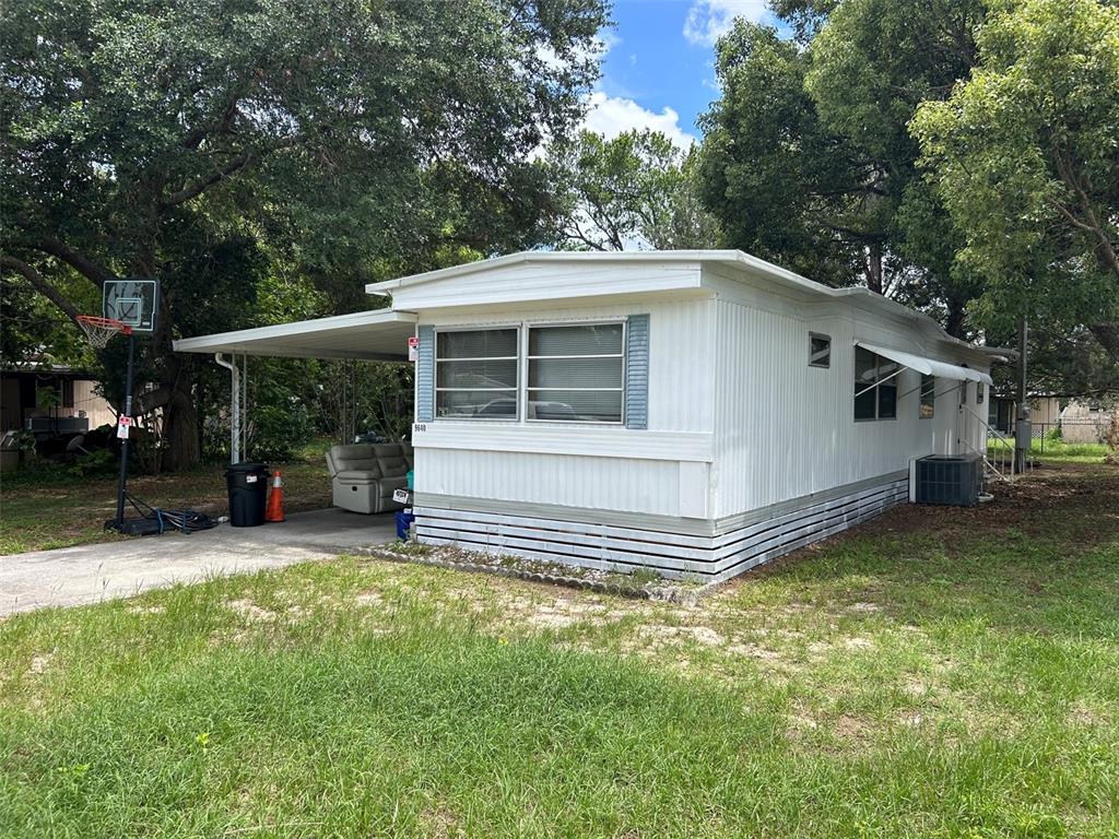 A white mobile home with a carport, basketball hoop, and surrounding trees on a grassy lot — 9640 SE 161ST PLACE, SUMMERFIELD, FL