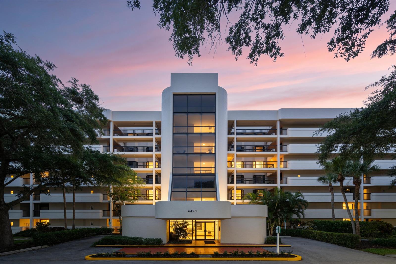 A multi-story apartment building with a central glass facade and curved entryway at dusk — 6420 Boca Del Mar Drive # 601, Boca Raton FL 33433, BOCA RATON, FL