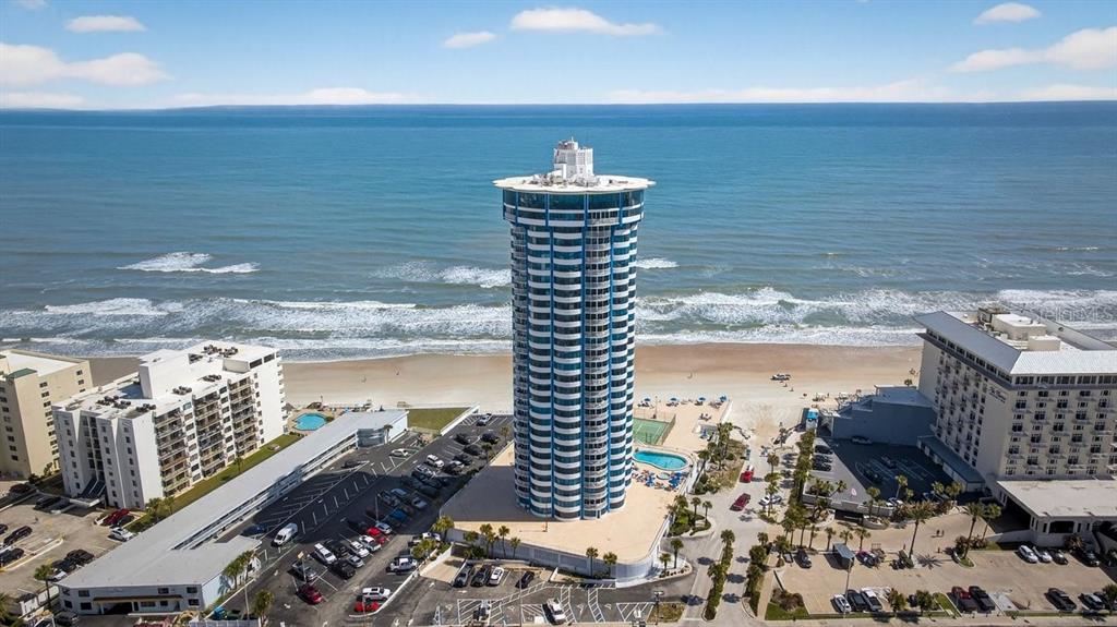 An aerial view shows a tall, cylindrical beachfront tower surrounded by parking lots and ocean — 2625 S ATLANTIC AVENUE UNIT 14NE, DAYTONA BEACH, FL