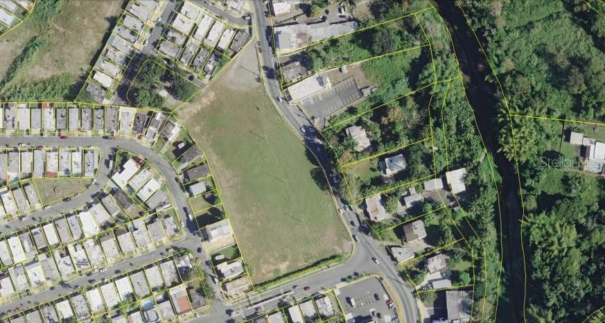 An aerial view shows a large, vacant grassy lot surrounded by residential buildings and roads — PR-174 SAN AGUSTIN, BAYAMON, PR