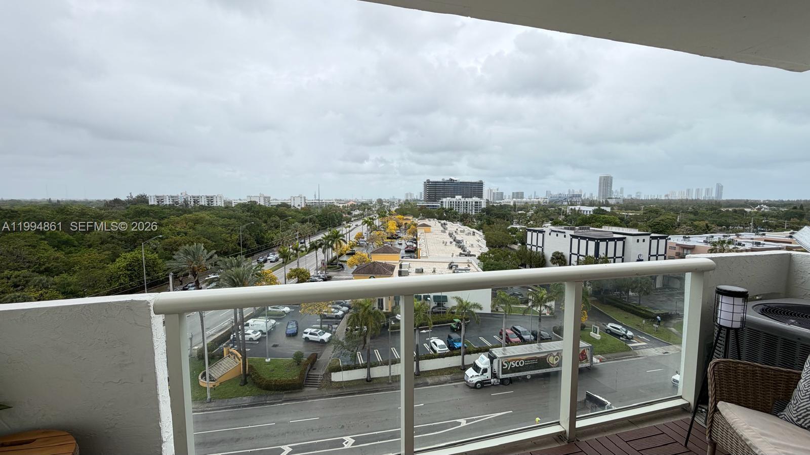A balcony overlooks a parking lot, a road with a Sysco truck, and distant city buildings — 13499 Biscayne Blvd # 805, North Miami FL 33181, NORTH MIAMI, FL