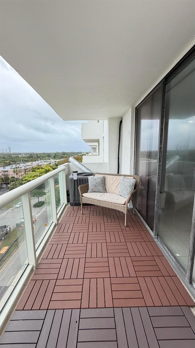 A balcony features interlocking wood-look deck tiles, a wicker loveseat, and a glass railing — 13499 Biscayne Blvd # 805, North Miami FL 33181, NORTH MIAMI, FL