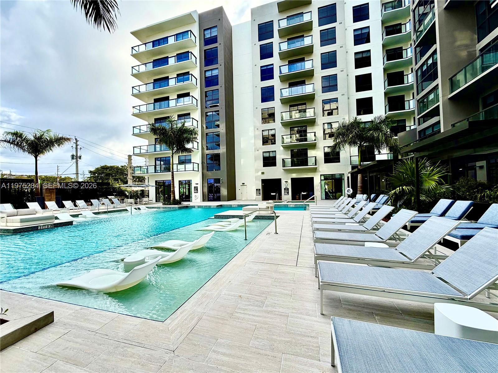 A modern apartment building features a swimming pool with submerged lounge chairs and poolside seating — 1650 NE 124th St # 621, North Miami FL 33181, NORTH MIAMI, FL