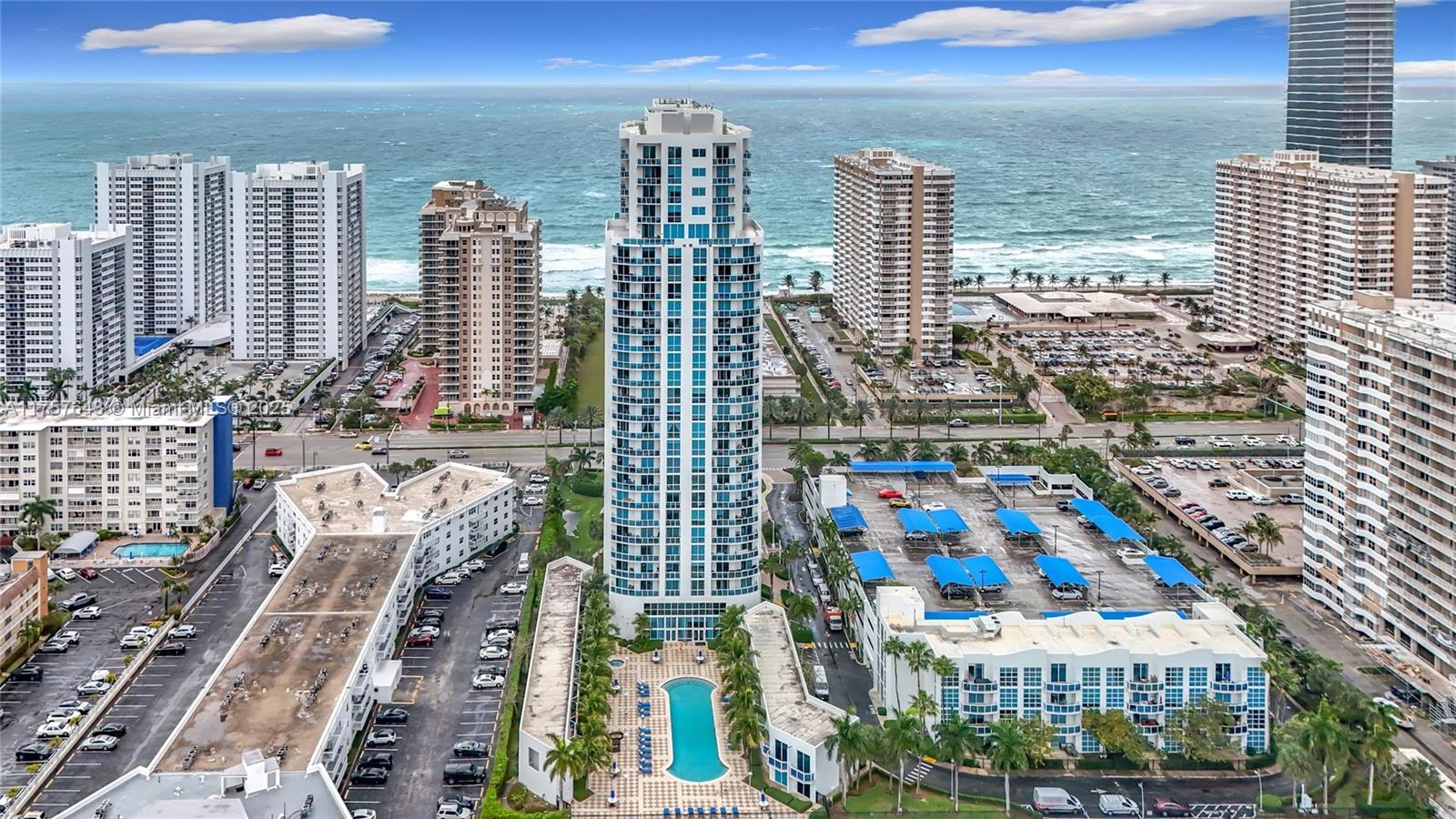 An aerial view shows a tall residential tower surrounded by parking lots and coastal buildings — 1945 S Ocean Dr # 605, Hallandale Beach FL 33009, HALLANDALE BEACH, FL
