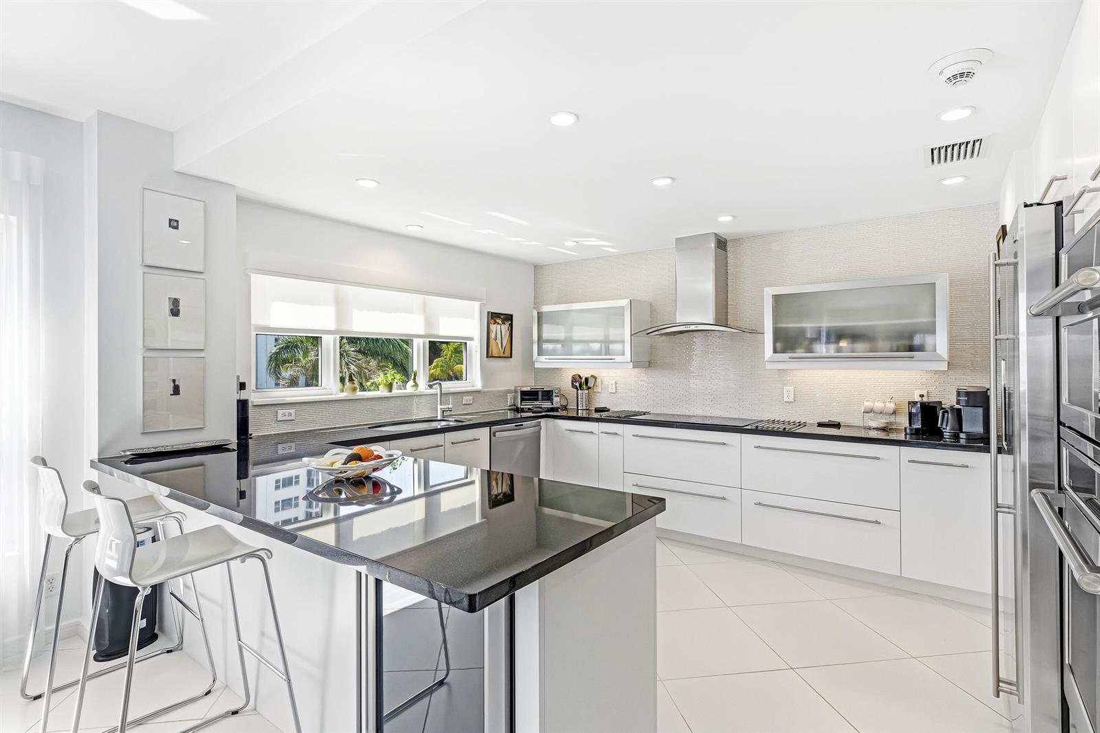 A modern kitchen features white cabinetry, black countertops, a breakfast bar, and stainless steel appliances — 200 Macfarlane Drive # 406, Delray Beach FL 33483, DELRAY BEACH, FL