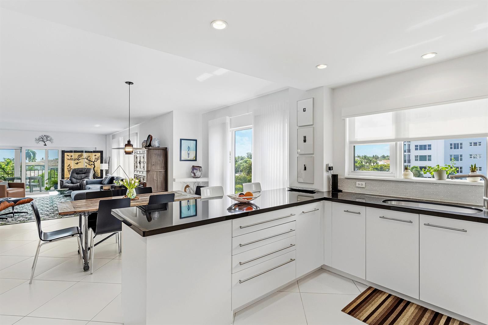 A modern kitchen with white cabinets and black countertops overlooks an open-concept dining and living area — 200 Macfarlane Drive # 406, Delray Beach FL 33483, DELRAY BEACH, FL