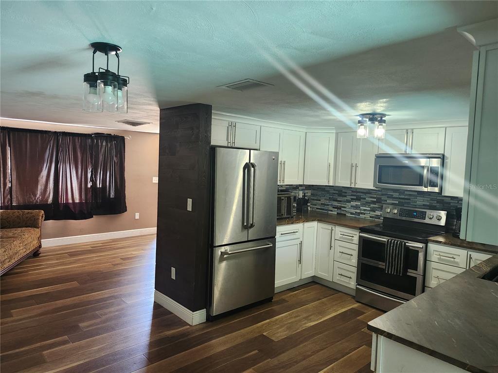 A kitchen with white cabinets, stainless steel appliances, and wood-look flooring opens to a living area — 8501 HUNTING SADDLE DRIVE, HUDSON, FL