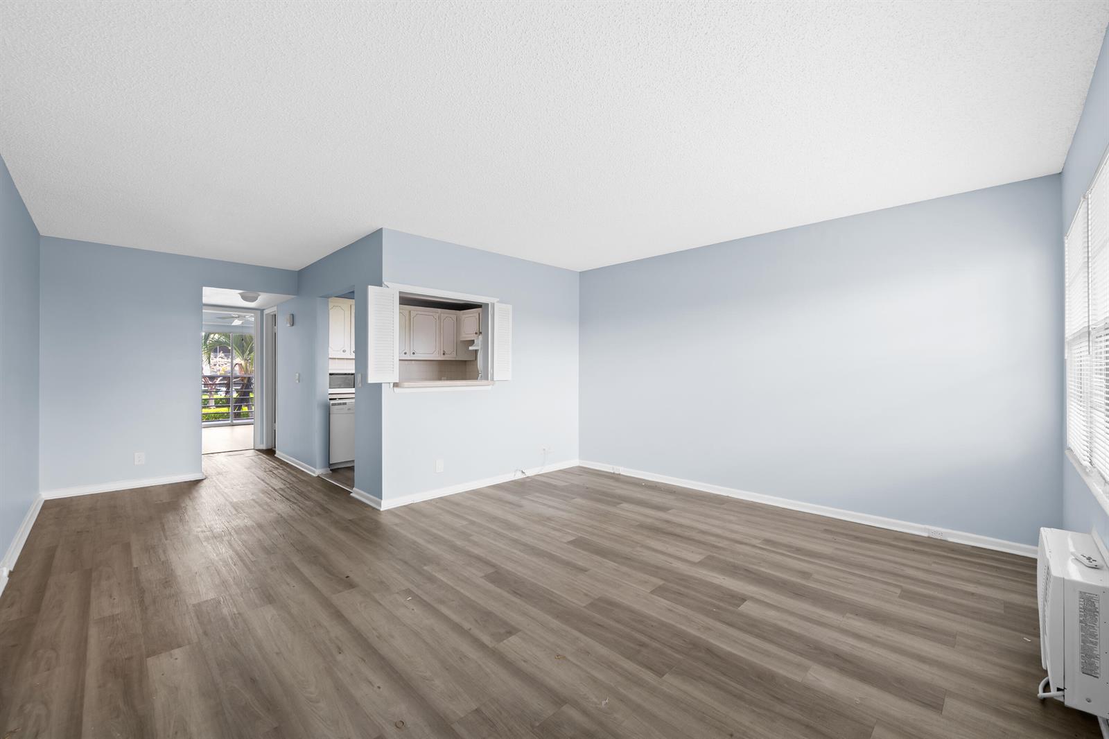 An empty living room with light blue walls, wood-look flooring, and a kitchen pass-through window — 148 Preston Way # 148, Boca Raton FL 33434, BOCA RATON, FL