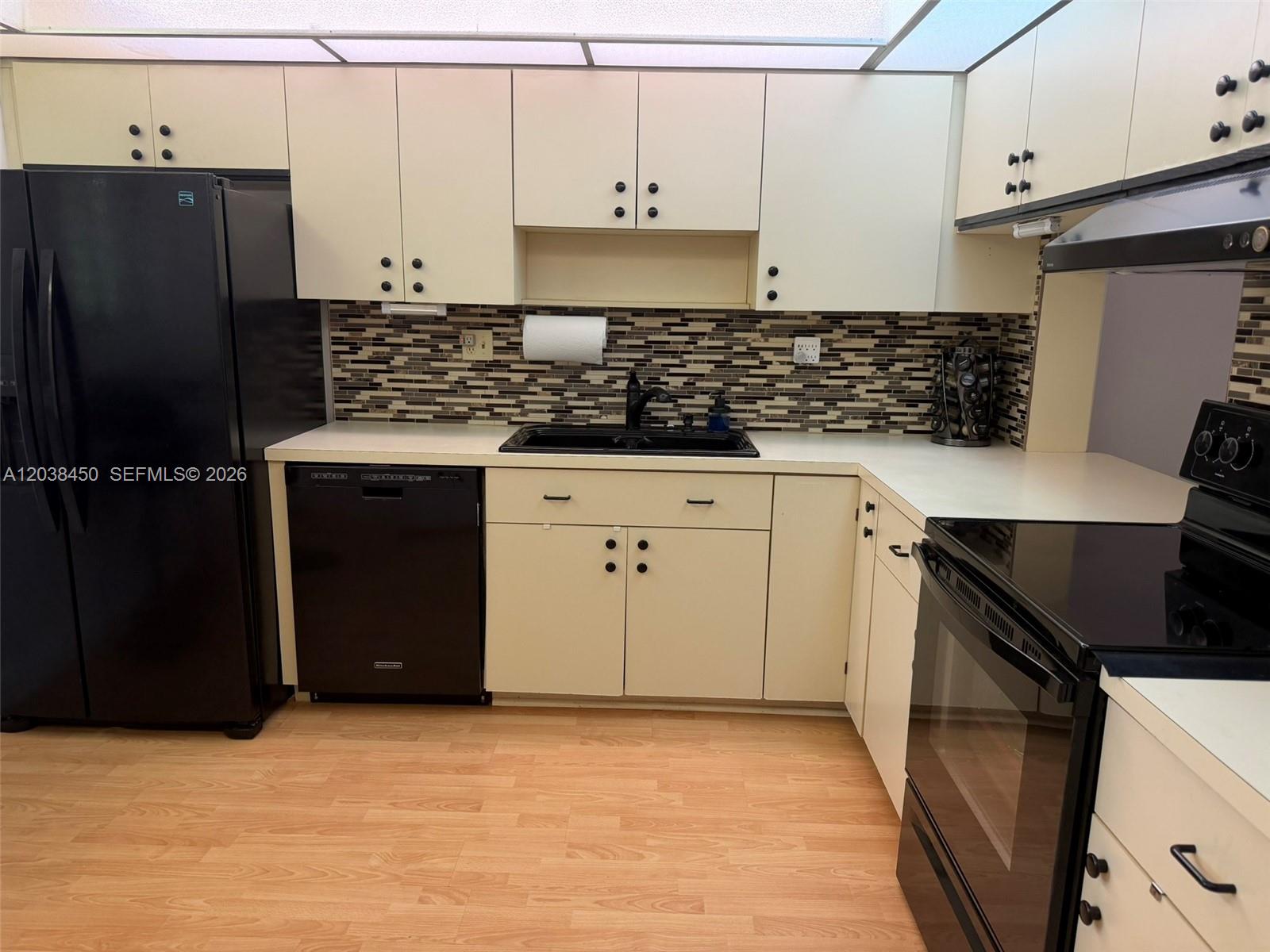 A kitchen features off-white cabinets, black appliances, a mosaic tile backsplash, and light wood flooring — 4960 E Sabal Palm Blvd # 308, Tamarac FL 33319, TAMARAC, FL