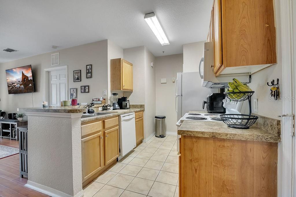 A kitchen with light wood cabinets, laminate countertops, and tile flooring opens to a living area — 5550 E MICHIGAN STREET UNIT 2114, ORLANDO, FL