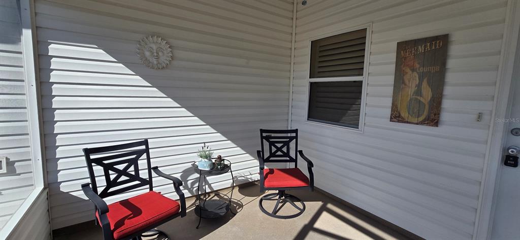 A covered porch features white vinyl siding, two chairs with red cushions, and a small table — 3489 CAMBRIA CIRCLE, THE VILLAGES, FL