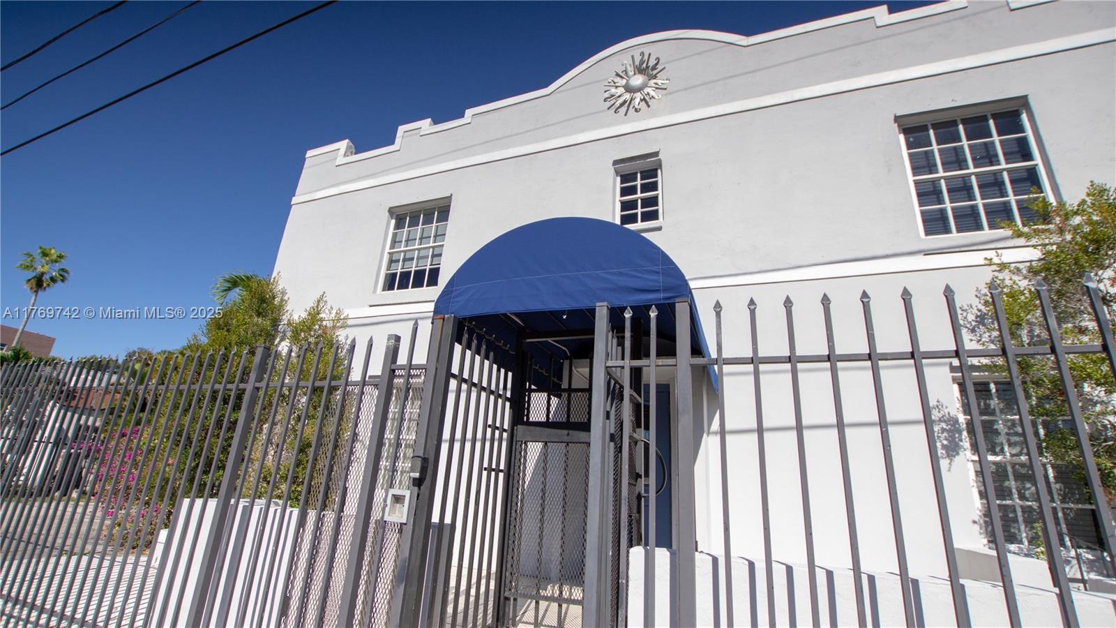 A low-angle view shows a light-colored building exterior with a blue awning and metal fence — 577 NE 62 STREET # 2, Miami FL 33138, MIAMI, FL