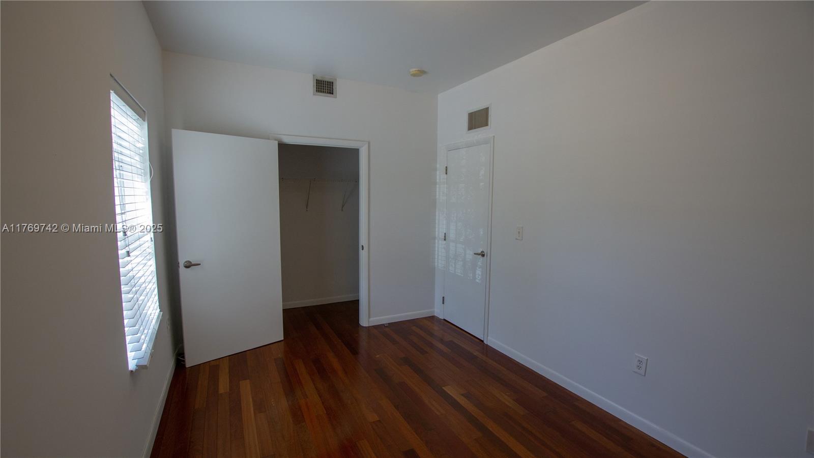 An empty bedroom features white walls, hardwood floors, a window with blinds, and two doors — 577 NE 62 STREET # 2, Miami FL 33138, MIAMI, FL