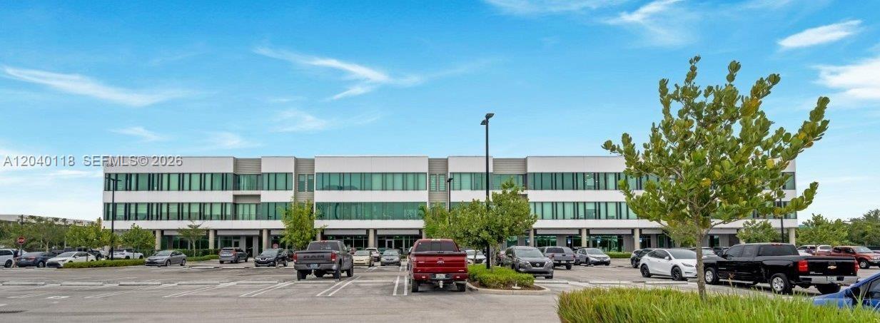 A three-story commercial office building stands behind a large parking lot with several vehicles — SWEETWATER, FL