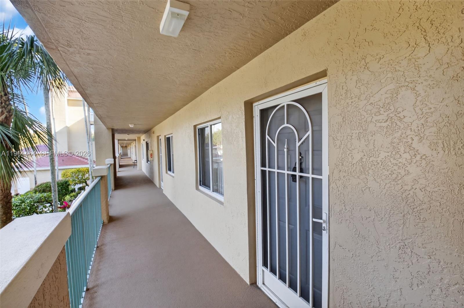 An outdoor apartment walkway features a beige stucco wall, a white security door, and railings — 12540 Majesty Cir # 208, Boynton Beach FL 33437, BOYNTON BEACH, FL