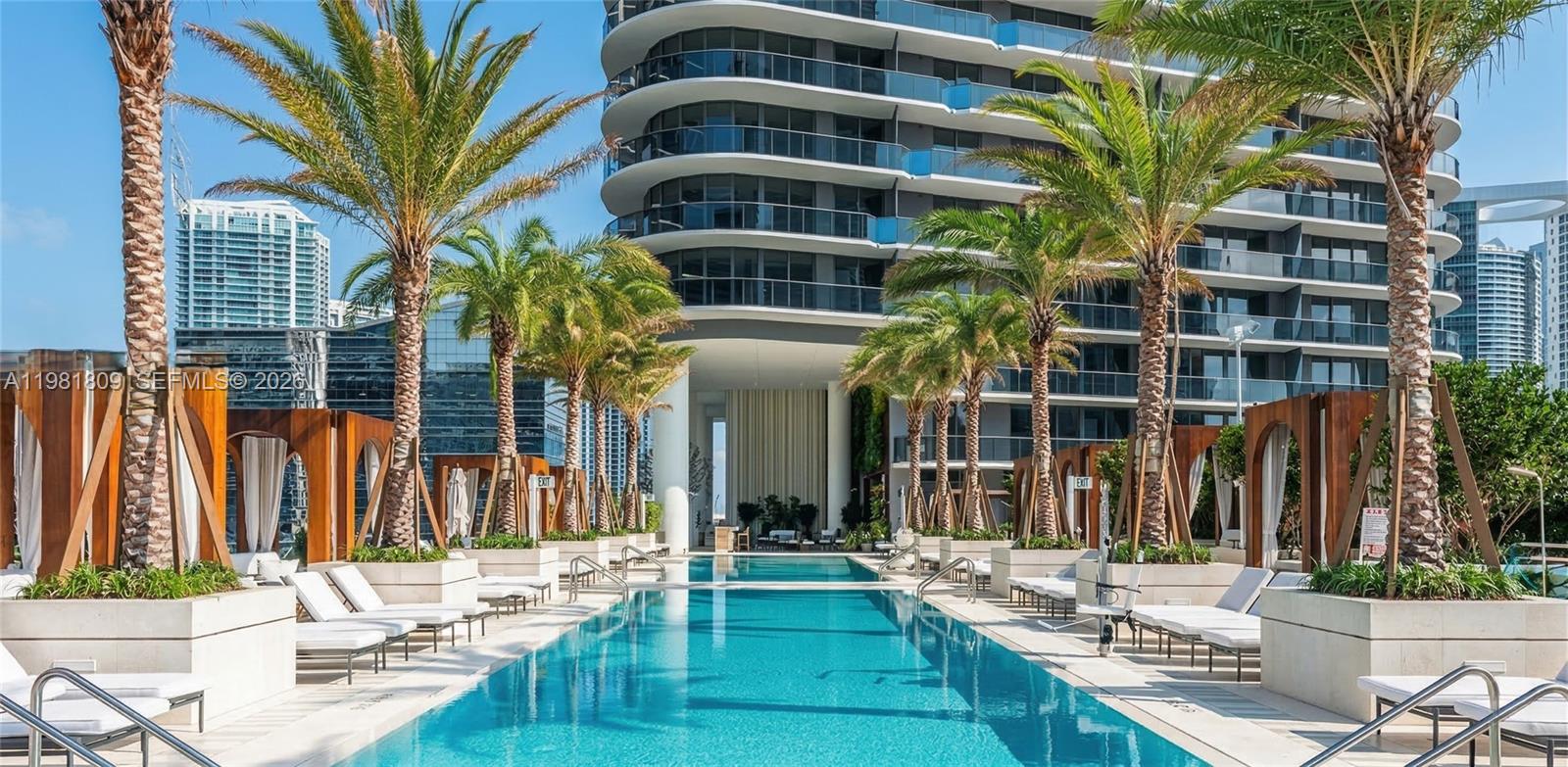 A long rectangular swimming pool is surrounded by lounge chairs, palm trees, and modern buildings — 801 S Miami Ave # 1203, Miami FL 33130, MIAMI, FL