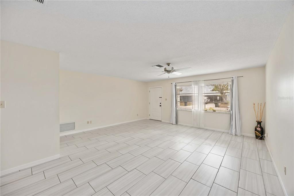 An empty living room features light-colored tile flooring, white walls, a ceiling fan, and windows — 181 SANDHURST DRIVE, VENICE, FL