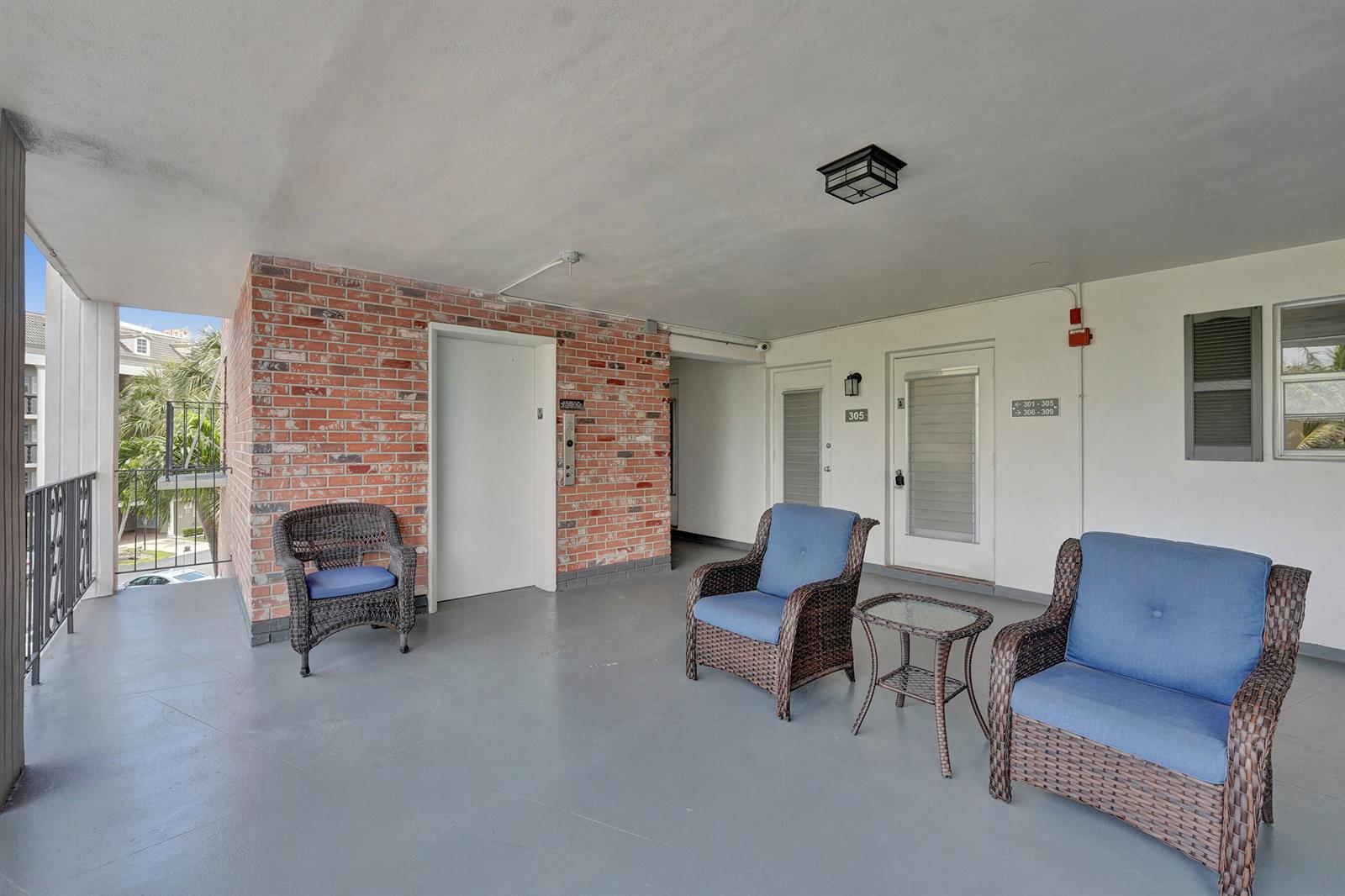 An outdoor covered walkway features a brick wall, wicker chairs, and apartment unit doors — 455 Paradise Isle Boulevard # 305, Hallandale Beach FL 33009, HALLANDALE BEACH, FL