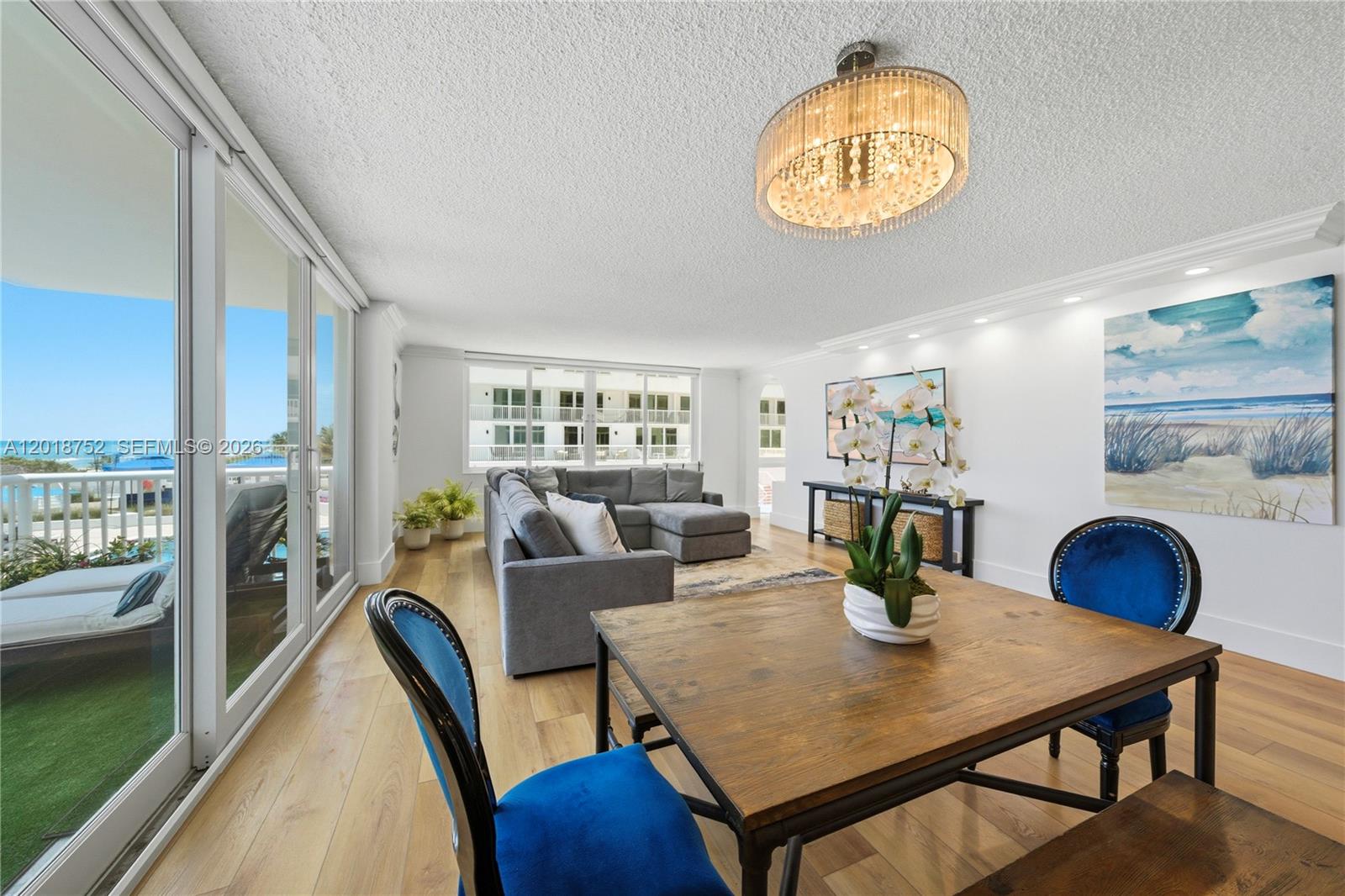 A bright living and dining area features wood floors, a crystal chandelier, and balcony views — 5401 Collins Ave # 102, Miami Beach FL 33140, MIAMI BEACH, FL