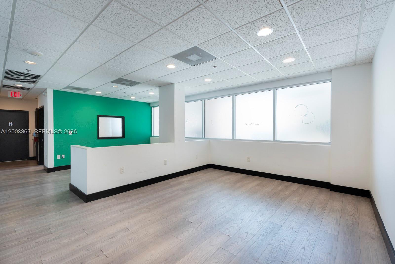 An empty office space features light wood flooring, a green accent wall, and large windows — 6141 Sunset Dr # 110, South Miami FL 33143, SOUTH MIAMI, FL