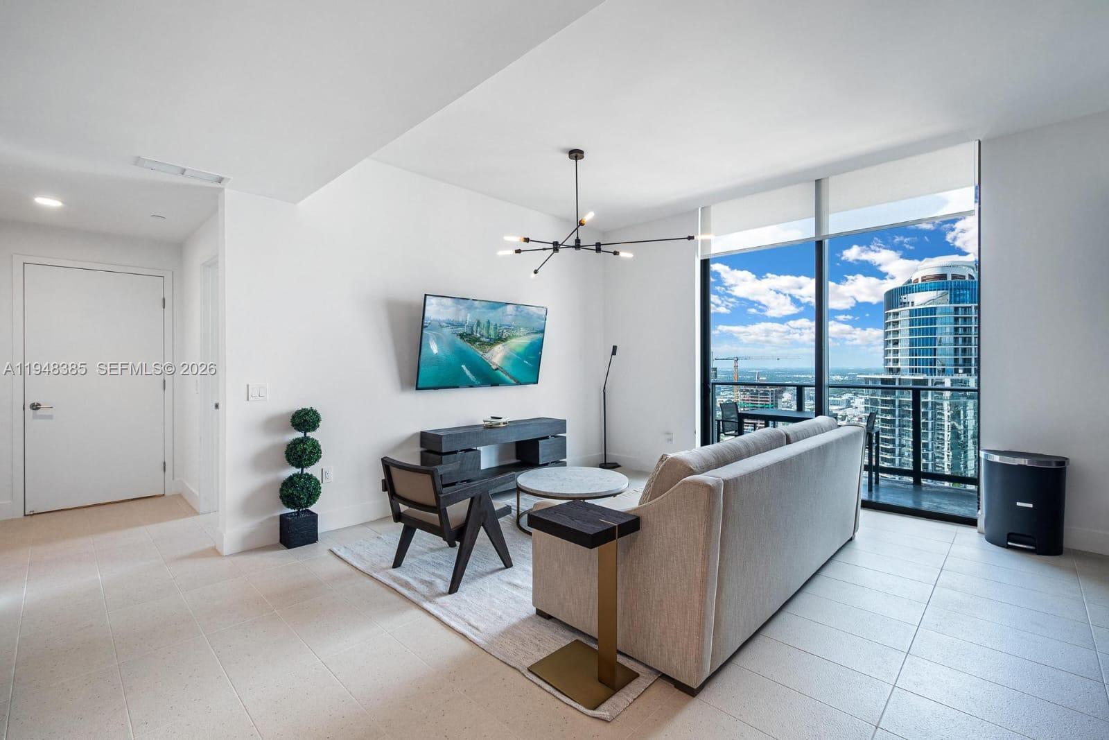 A modern living room features a sofa, television, and floor-to-ceiling windows with city views — 159 NE 6th St # 5101, Miami FL 33132, MIAMI, FL