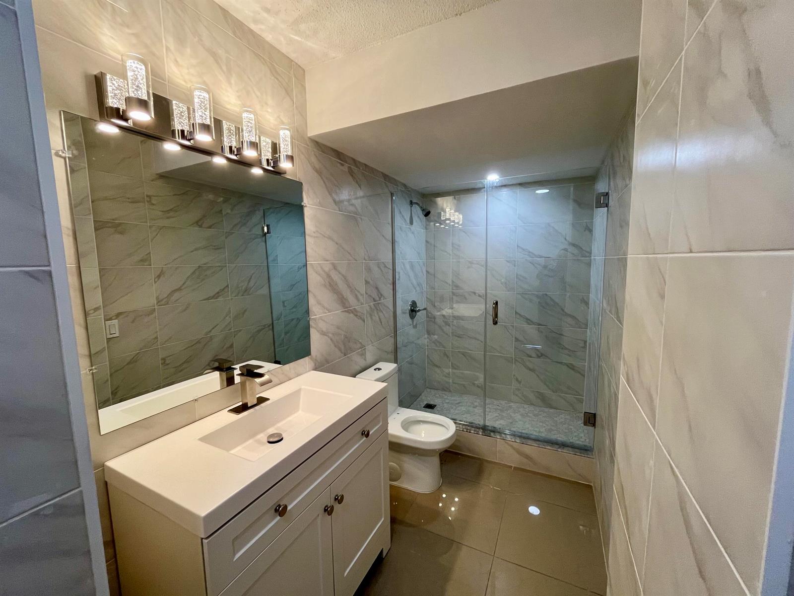 A modern bathroom features a white vanity, a glass-enclosed shower, and light-colored marble-style wall tiles — 1801 S Ocean Drive # 633, Hallandale Beach FL 33009, HALLANDALE BEACH, FL
