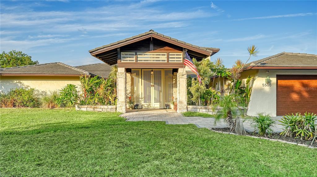 The exterior of a single-story house features a stone-pillared entryway and a wooden garage door — 5500 MERRIMAC DRIVE, SARASOTA, FL