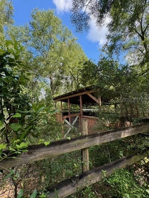A wooden fence stands in the foreground before a rustic, open-sided animal shelter in a forest — SW COLES COURT, FORT WHITE, FL