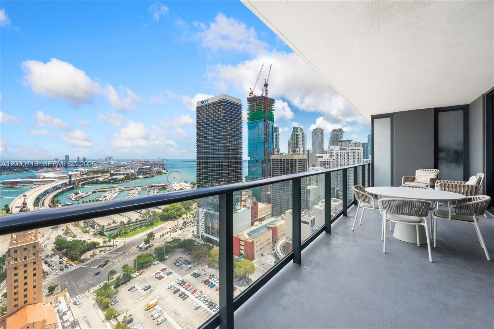 A high-rise balcony features outdoor dining furniture overlooking a city skyline and a harbor — 159 NE 6th St # 2915, Miami FL 33132, MIAMI, FL
