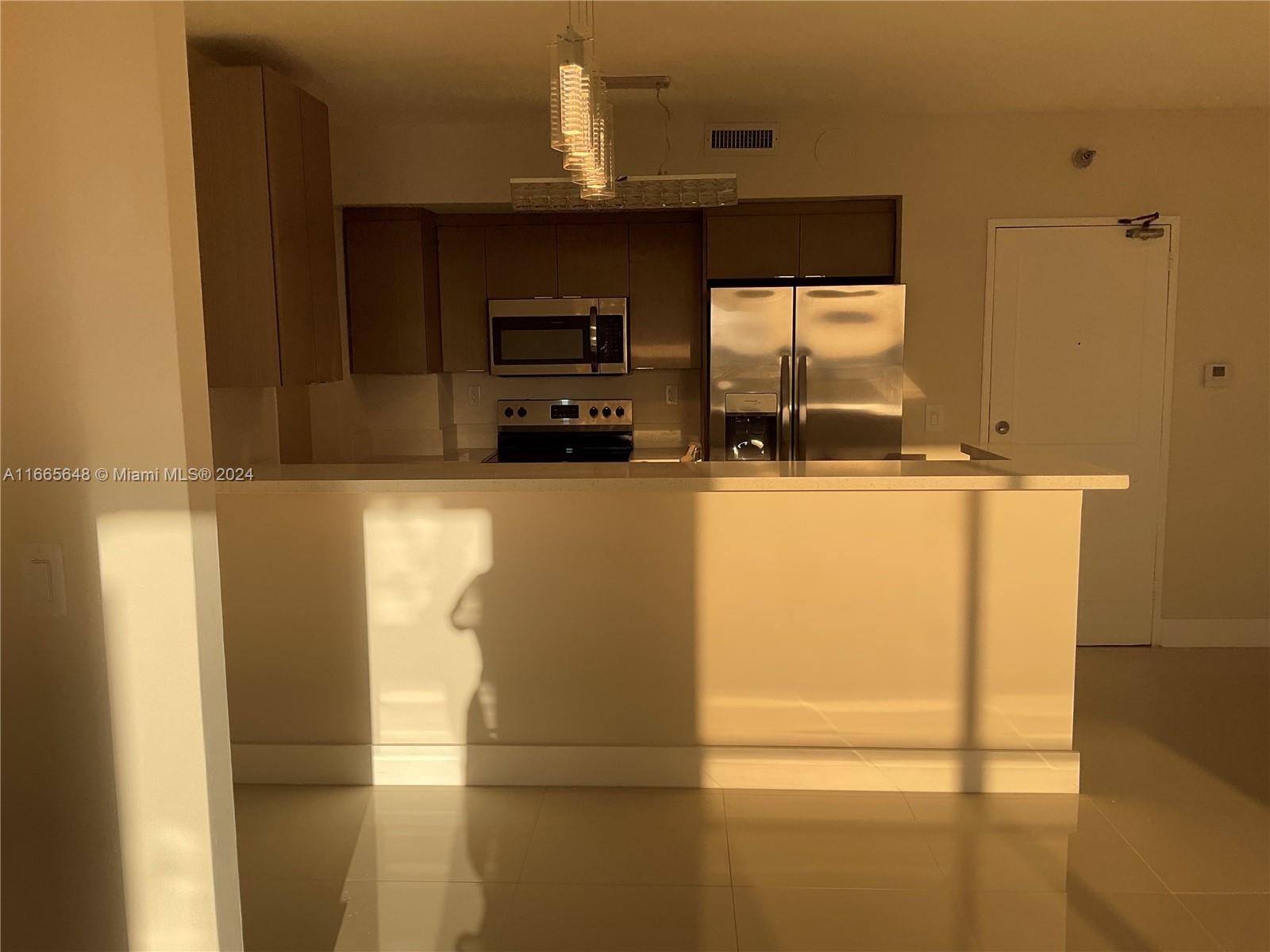 A kitchen features stainless steel appliances, brown cabinetry, and a breakfast bar with pendant lighting — 10185 Collins Ave # 406, Bal Harbour FL 33154, BAL HARBOUR, FL