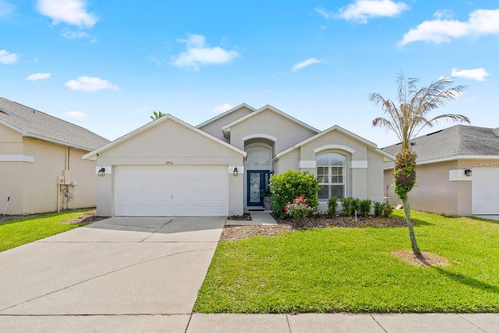 A single-story suburban house with a two-car garage, light stucco exterior, and a palm tree — 2916 BLOOMING ALAMANDA LOOP, KISSIMMEE, FL