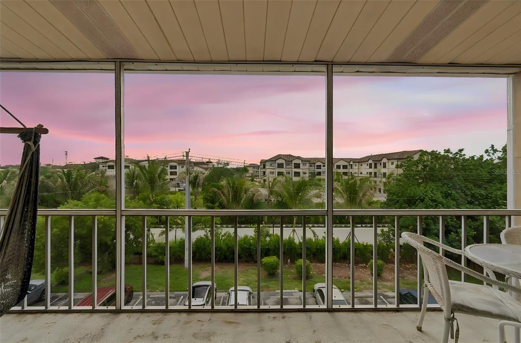 A screened-in balcony overlooks a parking lot, lush greenery, and apartment buildings at sunset — 1714 69TH AVENUE W UNIT C401, BRADENTON, FL