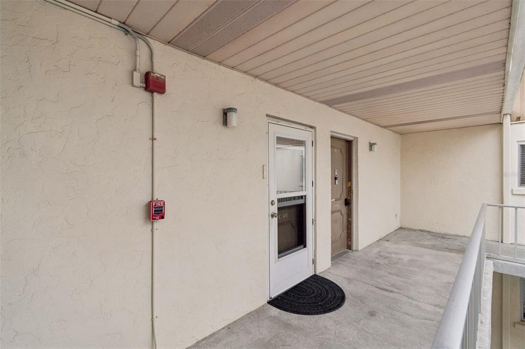 An outdoor apartment walkway features a white storm door, a brown entry door, and a fire alarm — 1714 69TH AVENUE W UNIT C401, BRADENTON, FL