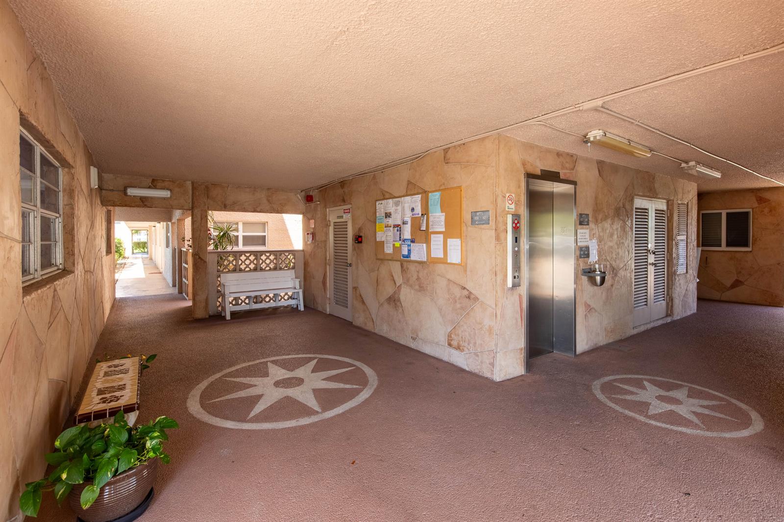 An outdoor apartment building lobby features stone walls, an elevator, a bulletin board, and benches — 2615 NE 1st Court # 411, Boynton Beach FL 33435, BOYNTON BEACH, FL